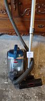 Full view of the Bissell PowerForce Cyclonic vacuum cleaner including the hose, extension wand, and floor brush head on a concrete floor with a wooden dresser in the background.
