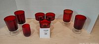 Seven cranberry glassware pieces arranged in a group on a light wood surface in front of a white backdrop. Includes four small tumblers, two medium tumblers, and one tall tumbler.