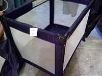 Side view showcasing mesh fabric sides and structure of fold-able baby play pen with carry bag to the left.