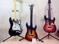 Three Hero guitars on stands: one black and white, one sunburst color, one red with stickers. One stand missing part.