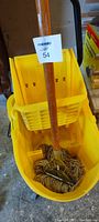 Photo show yellow industrial mop bucket with wringer, black handles, and mop inside with wooden handle and string mop head.