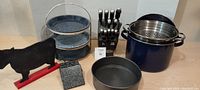 Kitchen items displayed including blue stock pot with stainless steel colander, pie plate set on two-tier stand, black knife block with knives, cow-shaped chalkboard, granite coasters.