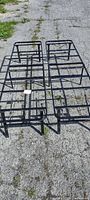 Photo showing folded black metal bed frame laid on pavement, displaying metal grid and foldable legs.
