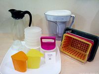 Overall view of miscellaneous plastic kitchen items including water jugs, baskets, patty maker, containers, and tray on a flat surface.