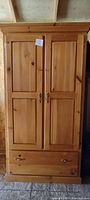 Front view of closed pine colonial style armoire showing two paneled doors and bottom drawer with metal handles.