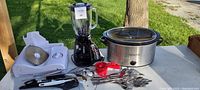 Wide view of all items in the lot including slow cooker, blender, meat slicer, and utensils on table outdoors