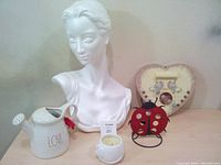 A photo showing a white female bust sculpture, ceramic 'LOVE' pitcher, small candle holder, red ladybug decoration, and heart-shaped photo frame.