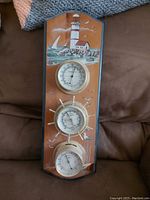 Wall-mounted wooden plaque with a nautical lighthouse painting and three round instruments: barometer, thermometer, and hygrometer in ship's wheel design.