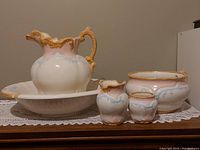 Photo showing the entire antique ceramic wash basin set including large basin, water pitcher, chamber pot, and two smaller containers on a lace doily.