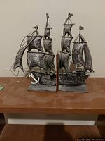 Pair of metal ship bookends on wooden surface against plain wall, showing detailed sails, rigging, and anchor accent on one bookend.