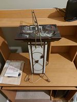 Photo of vintage candy tray or ashtray on wooden desk, showing rectangular metal tray with leaf design, mounted on a decorative tall stand with white inserts and metal frame. Includes visible power cord for plug-in illumination.
