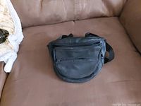 Photo of a small black pleather fanny pack laid on a brown couch, showing the front with a main zipper and smaller front zipper compartment.