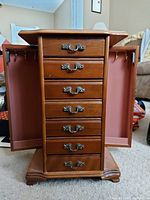 Front view of the wooden jewelry box with side doors open showing hooks for necklaces and six drawers with metal handles.