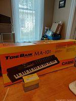 Casio ToneBank MA-101 keyboard in original orange box placed on a surface by a window with natural lighting. Box intact with product images and branding visible.