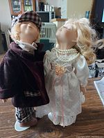 Photo of two porcelain dolls posed kissing, dressed in formal vintage clothing, male doll in burgundy velvet coat and tartan pants, female doll in white satin dress with pearls and lace.