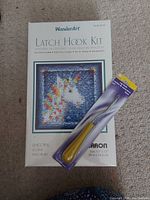 Photo shows WonderArt Unicorn Latch Hook Kit box and a packaged yellow-handled latch hook tool on carpet.