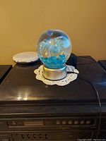 Clear glass decorative water globe with three blue dolphins inside, mounted on a silver base with light and motion mechanism. Globe resting on a white crocheted doily on a black surface against a beige wall.