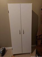 White pantry cabinet with closed double doors, silver handles, standing against wall on carpeted floor.