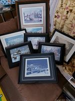 Photo showing eight framed prints arranged on a brown chair and adjacent space, varying frame colors black and wood tone, depicting winter and rural scenes from Sparta Ontario.