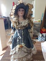 Single porcelain Victorian doll wearing a detailed blue dress, lace accents, curled hair, and a large feathered hat standing upright in room.