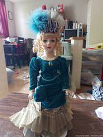 Full frontal image of a Victorian style porcelain doll wearing a green velvet dress with puffed sleeves and layered skirt with gold pleats, holding a matching clutch and wearing a feathered decorative hat.