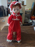 Porcelain doll with red hair, red dress with white lace, bonnet, slippers, and teddy bear accessory.
