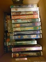 Photo of 14 VHS tapes stacked in a box, titles visible include Disney classics and other children's movies and shows.