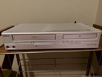 Front view of white Symphonic VHS/DVD player showing VHS slot on left and DVD tray on right with various control buttons and ports.