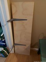 Photo showing a wooden plywood shelf measuring approximately 48 by 16 inches leaning against a wall, with two metal wall-mount brackets attached to the underside of the shelf.