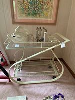 Side angle view of the cream metal and glass bar cart with two shelves and bottle holders, with figurines on top (not included).
