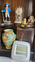 Display shelf showing soldier figurine, anniversary clock under glass dome, ceramic dog, resin elephant, and part of handmade urn vase, and Sharp digital clock below.