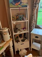 White ladder style display stand shown with various decorative items on shelves (not included in sale). The stand has three open shelves and one drawer below. The stand leans against a wall beside a window and small side table.