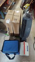 Lot showing two boxed Holmes window fans, one gray Lasko round fan standing upright, and two small coolers on the floor.