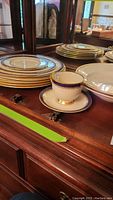 Group view of stacked Lenox Eternal plates with gold trim and Jefferson teacup with blue and gold detailing on wooden surface.