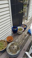 Outdoor photo showing multiple ceramic planters, a metal plant stand with a blue planter containing a small tree, terracotta and beige pots, and two round decorative ceramic trays with soil, placed on a wooden deck.