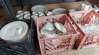 A collection of white dishes including stacked plates, cups, and pink floral patterned serving dishes in crates.
