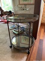 Front and side view of the two tiered metal serving table with mirrored surfaces and wheels on a carpeted floor near a doorway.