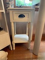 Front angled view of small white nightstand showing drawer and open shelf below, positioned next to window and other furniture.