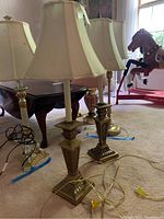 Two brass Stiffel lamps with cream-colored fabric shades standing on carpet. Lamps show some pitting and tarnish on bases. Electrical cords with yellow plugs visible.
