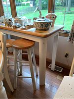 Counter height table with butcher block top and white painted legs, accompanied by two wooden stools with white legs, photographed near window with dishes on top.