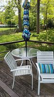 Photo of round glass top table with black metal base, accompanied by blue and white striped umbrella and four white plastic chairs on wooden deck.