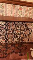 Full view of the metal base with swirled circular wine bottle holders and attached wooden top surface against floral wallpaper.