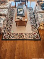 Full view of the area rug placed between two floral patterned sofas with a wooden coffee table on it showing its overall size, floral border pattern and beige center.