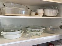 Glass casserole dishes and bowls arranged on kitchen shelving, various styles including vintage Pyrex with green floral pattern, clear and white opaque glass dishes, some with lids.