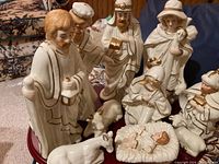 Close-up photo of ceramic figures including Mary, Joseph, baby Jesus in a manger, three wise men, shepherd, angel, and three animal figurines arranged on a carpet.