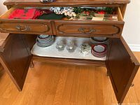 Wide view of vintage wooden sideboard with drawer open showing Christmas-themed dishes and stacked plates below, cups and small bowls.