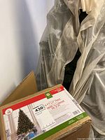 Photo showing assembled artificial Christmas tree partially behind protective fabric with its original box labeled '15' and detailed product description including height and lighting.