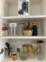 Assortment of measuring cups and kitchen containers including jars, pepper grinder, and glassware on shelf