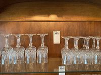 Sixteen crystal wine glasses displayed in a wooden cabinet shelf. Eight glasses on the left have larger bowls (red wine), eight on the right have smaller bowls (white wine).