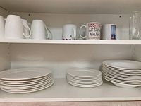 Top shelf with eight white textured design mugs, three assorted mugs with printed designs, and a tall clear glass tumbler